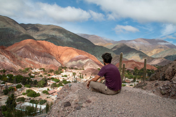 Hombre sentado en las montañas, mirando el pueblo de Purmamarca desde las alturas con los cerros coloridos de fondo.