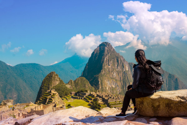 Mujer turista sentada en una piedra con vistas panorámicas hacia las montañas y parte de la ciudadela del sitio arqueológico de Machu Picchu.