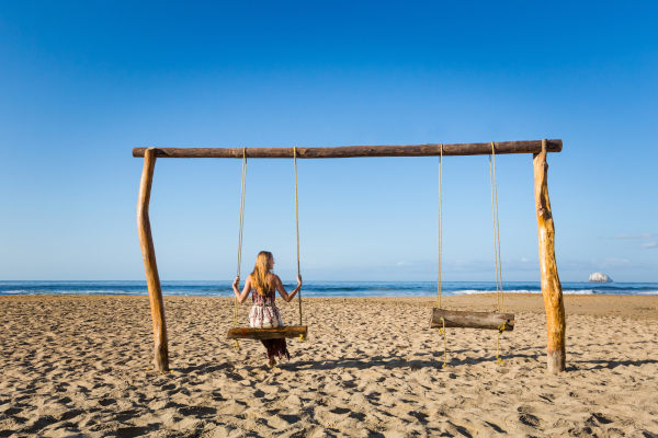 Mujer rubia turista con vestido de playa sentada en un columpio en Playa Zipolite, México, con vistas hacia el mar.