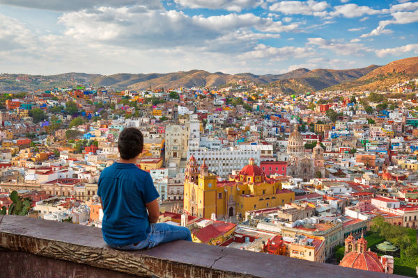 Turista masculino sentado en una barandilla de un mirador con vistas a la ciudad de Guanajuato, en México, en un día soleado.