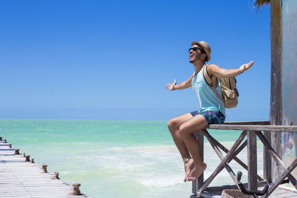 Hombre joven con mochila, sombrero, gafas de sol y traje de baño sentado en una barandilla de madera cerca de un muelle con el mar de fondo en México.