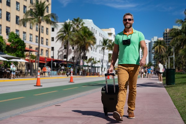 Un turista masculino caminando por las calles de Miami con una maleta y cámara de fotos.