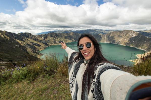 Joven viajera con poncho andino tomándose una selfie frente a la pintoresca laguna Quilotoa en los Andes ecuatorianos.