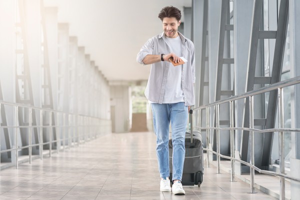 Hombre feliz comprueba la hora en su reloj de pulsera mientras camina con la maleta en el aeropuerto hacia la zona de embarque.