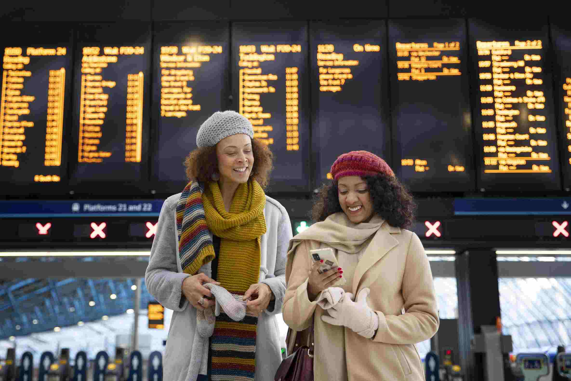 Duas mulheres felizes em aeroporto checam celular com painel de voos ao fundo, em foto que ilustra artigo sobre o que é milhas em viagens aéreas.