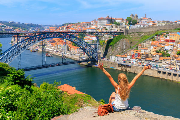 Mujer turista rubia con los brazos levantados sentada en un punto panorámico de Oporto con vistas al puente de hierro y la ciudad vieja.