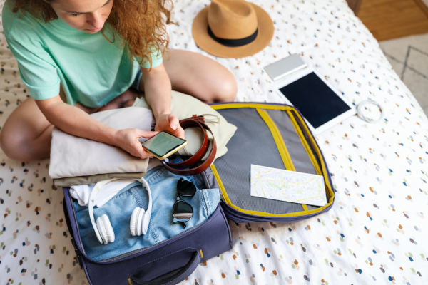 Mujer sentada sobre la cama, mirando el celular, con una maleta abierta recién preparada.