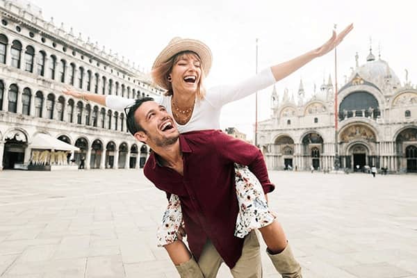 casal formado por homem e mulher juntos, em viagem para casal. Mulher está de chapéu, nas costas do homem