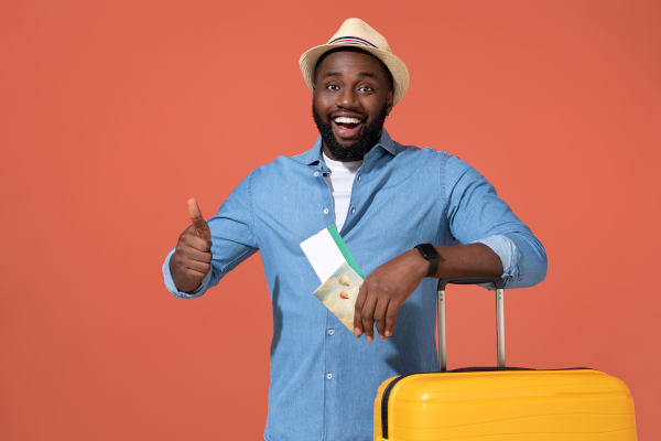Hombre con sombrero y camisa azul con pasajes aéreos en la mano y maleta listo para viajar con una sonrisa sobre un fondo liso.
