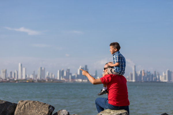 Padre sosteniendo en hombros a su hijo sentado en una roca señalando algo con la Ciudad de Panamá en el horizonte.
