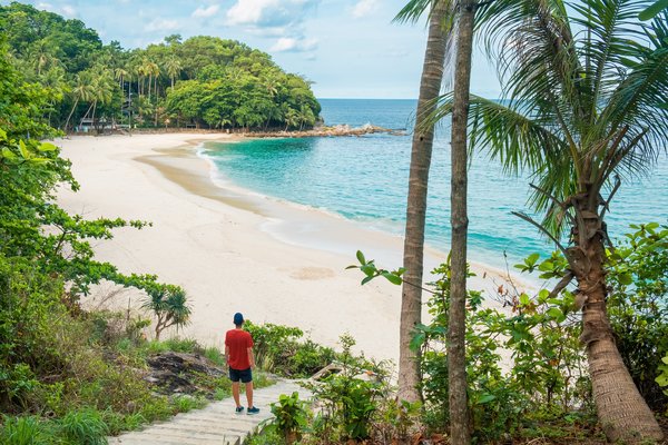 Turista masculino bajando una escalinata de piedra que conduce a una playa de arena tropical con el océano de agua turquesa en la isla de Phuket, Tailandia.