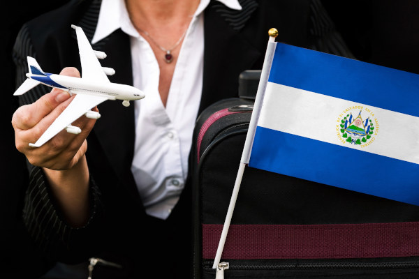 Mujer de negocios con un avión de juguete en la mano y una maleta con la bandera de El Salvador saliendo de uno de sus bolsillos con cierre.