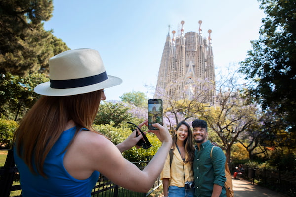  imagem de uma mulher vestida de azul, com chapéu, fotografando um casal em frente ao Templo Expiatório da Sagrada Família em Barcelona, Espanha, em viagem pela Europa.