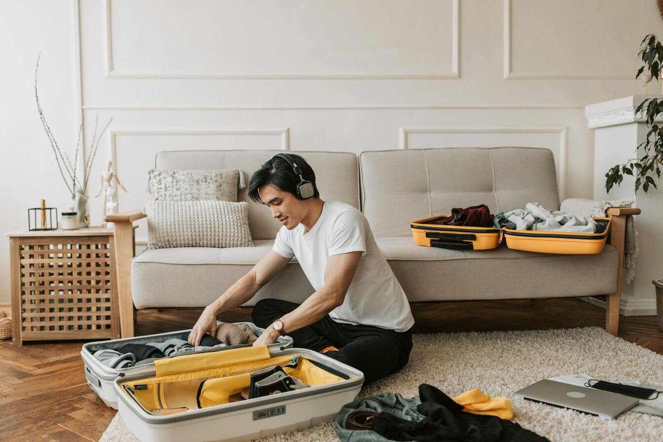  Homem organizando malas de viagem no chão da sala, com outra mala aberta sobre o sofá, representando o planejamento de férias 2025.