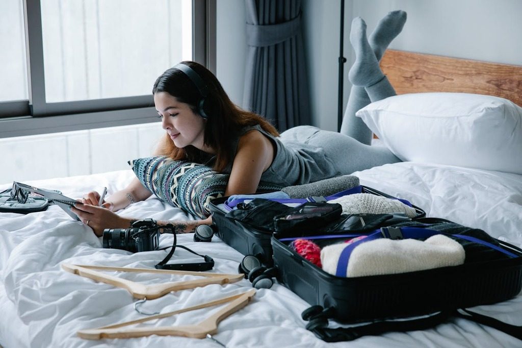 Mulher deitada na cama planejando viagem com mala aberta, câmera e roupas ao lado, representando o planejamento de férias 2025.