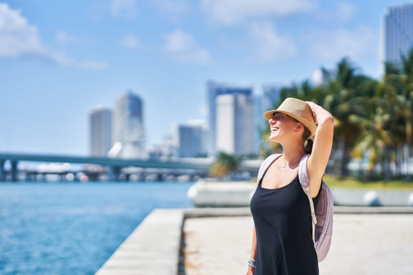 Mujer sonriente con sombrero mirando el mar con un fondo urbano soleado con palmeras en Miami, Florida, Estados Unidos