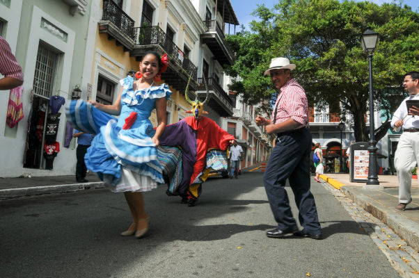 Muchacha con vestido celeste bailando en una calle de San Juan, Puerto Rico, acompañada por un hombre sonriente y una persona disfrazada.