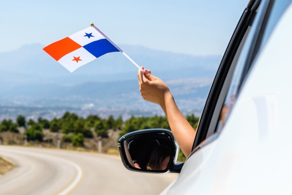 Persona ondeando pequeña bandera de Panamá por la ventanilla de un auto en movimiento en una carretera durante un día soleado.