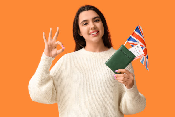 Mujer sonriente con suéter blanco, haciendo gesto de ok y sosteniendo pasaporte, boletos y bandera del Reino Unido sobre fondo naranja.