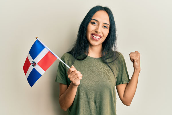 Mujer sonriente sosteniendo bandera de República Dominicana con una sonrisa en un fondo claro liso.