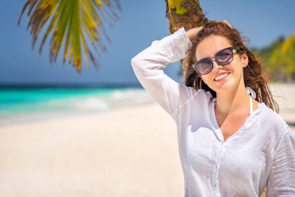 Mujer sonriente con camisa blanca y gafas de sol con la mano en la cabeza y la espalda apoyada en una palmera en una playa de arena blanca y aguas cristalinas en República Dominicana.