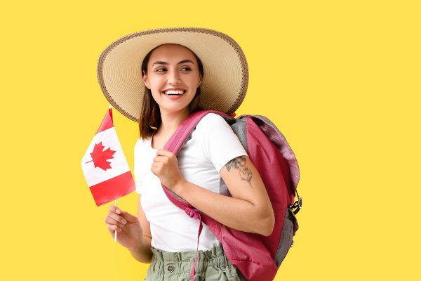 Mujer turista con mochila y sombrero sosteniendo bandera de Canadá con una sonrisa en fondo liso amarillo