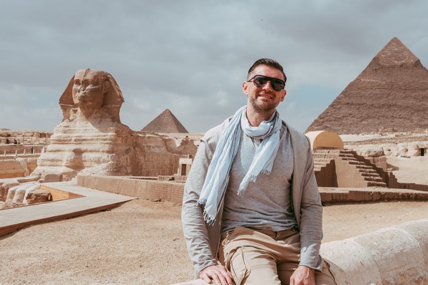 Hombre turista con gafas de sol sonriendo a la cámara con la Efigie y las Pirámides de Giza de fondo en un día nublado en Egipto
