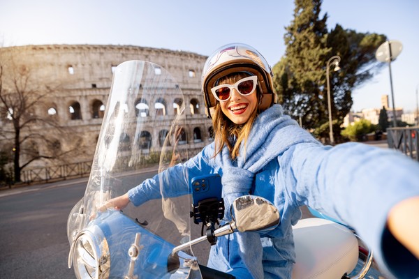 Mujer con casco y gafas de sol tomándose una selfie sobre un scooter que pasa por delante del Coliseo Romano en Roma, Italia.