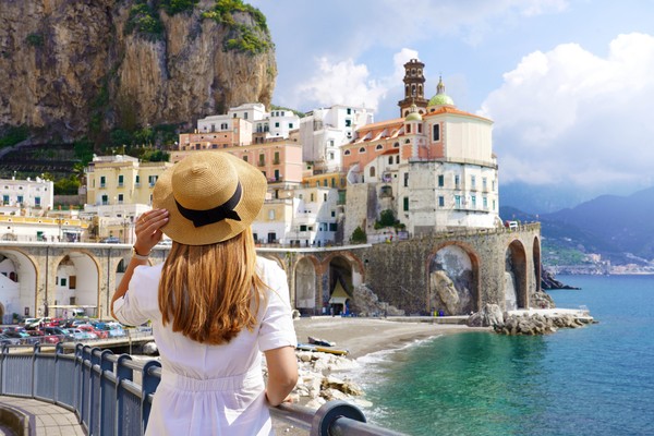 Vista posterior de una mujer sobre una barandilla con vistas hacia la costa con una pequeña playa en el pueblo de Atrani, Italia.