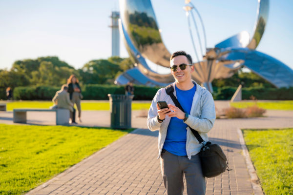 Turista usando su celular mientras recorre los parques de Buenos Aires, con la escultura Floralis Genérica al fondo.