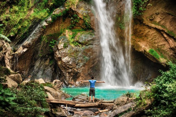 Joven parado con los brazos abiertos en frente de una cascada con un río celeste debajo y vegetación.