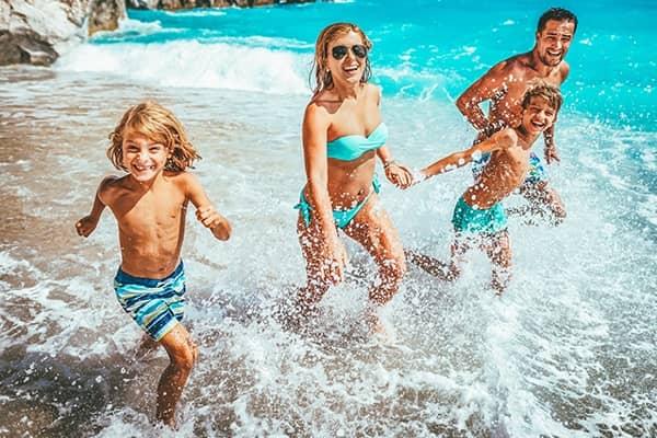 Família composta por mãe, pai, e dois meninos, correndo felizes na praia.