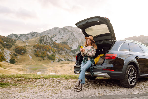Una joven viajera se sienta en la parte trasera de su automóvil, sonriendo y usando su teléfono en una pintoresca zona montañosa, con nubes colgando sobre las colinas.