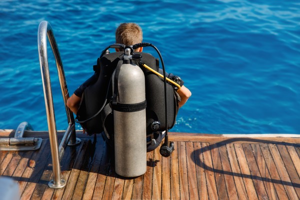 Hombre en traje de buceo sentado en la cubierta de madera de un yate con un tanque de oxígeno en la espalda preparándose para la inmersión.