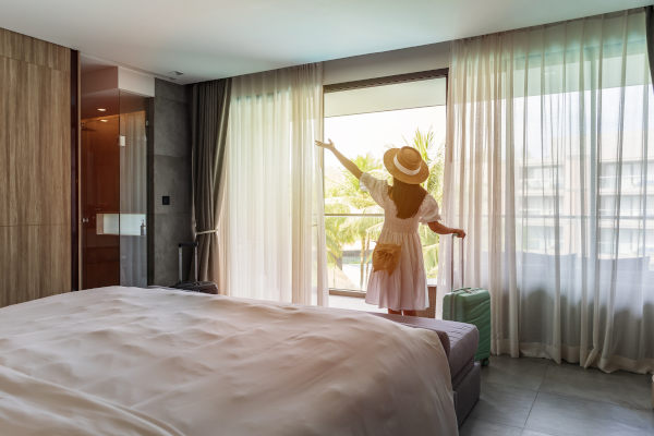Mujer joven con una valija mirando las vistas desde la ventana de una habitación de hotel.