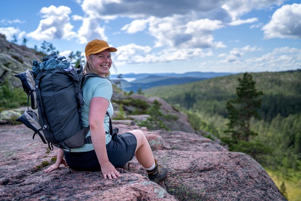 Mujer turista sentada en una roca con ropa de senderismo y mochila, sonriendo y mirando la cámara, con vistas a las montañas del parque nacional Skuleskogen, Suecia.