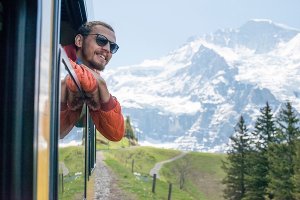 Hombre con gafas de sol asomado por la ventanilla de un tren con destino a Jungfrau, Suiza, con pinos y montañas nevadas de fondo