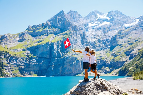 Dos hermanos ondeando bandera suiza parados sobre una roca en las orillas del lago Oeschinen, Alpes suizos.