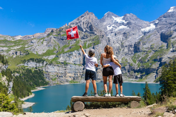 Familia feliz parada en un banco de troncos en un mirador con vistas al lago Oeschinen y la montaña de los Alpes, Suiza, en un día soleado y cálido.