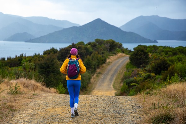 Mujer senderista caminando por un camino en la costa de Picton, Nueva Zelanda, con colinas verdes de fondo