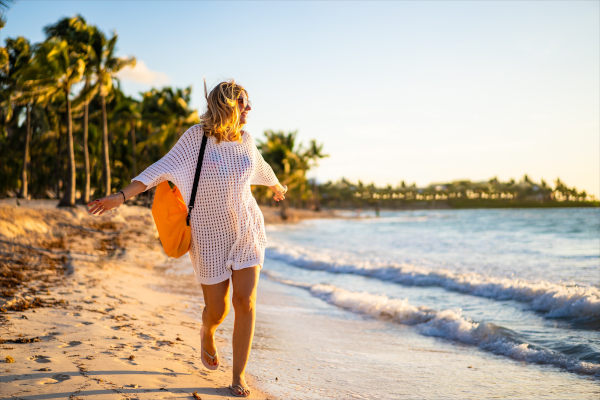 Mujer turista con bolso caminando con los brazos abiertos y una sonrisa en una playa tropical soleada al atardecer.