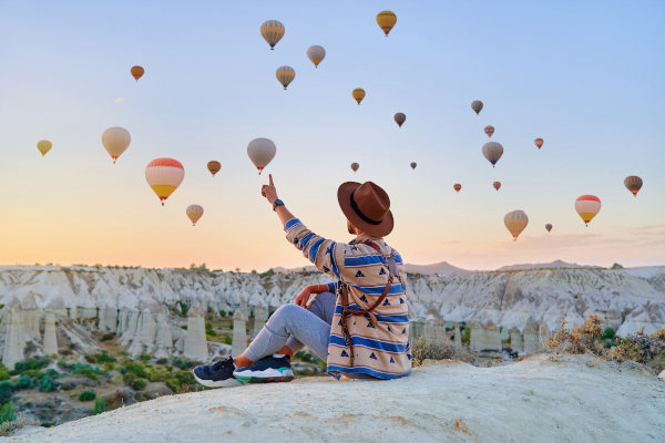 Turista sentado en una formación rocosa señalando los globos aerostáticos que sobrevuelan el valle de Goreme, Turquía, con las famosas 