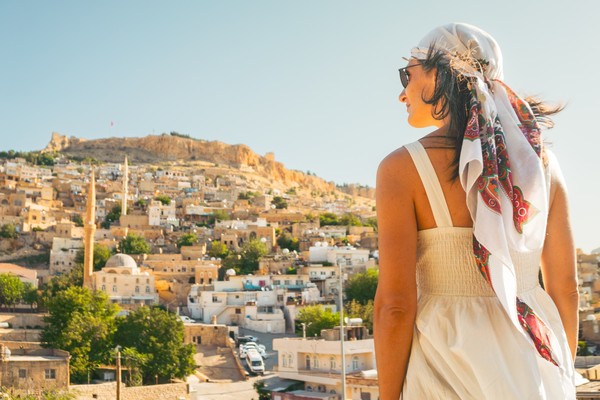 Mujer viajera con vestido blanco mirando la ciudad histórica de Mardin, Turquía, con minaretes de mezquitas, casas tradicionales y el castillo de Mardin en la cima de la colina