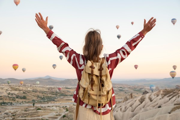 Mujer viajera con mochila alzando los brazos de espalda a la cámara y frente a los globos aerostáticos de Capadocia al amanecer, Turquía