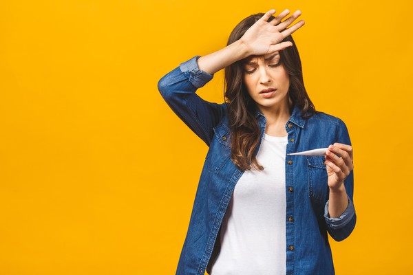 Mujer tomándose la cabeza mirando un termómetro con fondo amarillo liso.