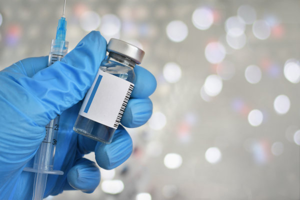 Mano con guante de látex de profesional de la salud sosteniendo jeringa con aguja y con un refuerzo genérico de vacuna en un vial con etiqueta en blanco.