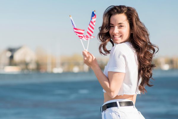 imagem de mulher vestida com uma camiseta branca segurando na mão esquerda uma bandeira dos EUA representando viagem para os estados unidos