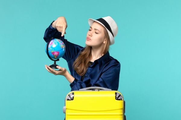 mulher de chapéu, apoiada em uma mala de viagem amarela, apontando para um globo terrestre em miniatura representando viagem para Europa