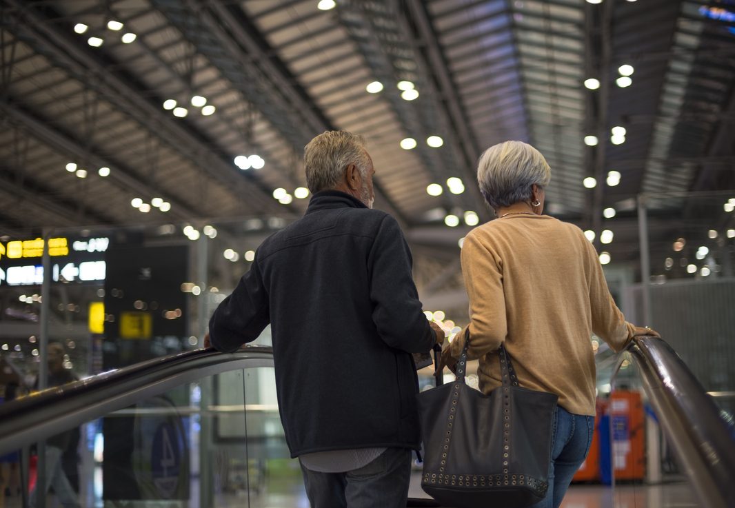 Casal de idosos em aeroporto internacional, subindo escada rolante com bagagem de mão, simbolizando a importância de escolher seguro viagem internacional qual o melhor para garantir tranquilidade