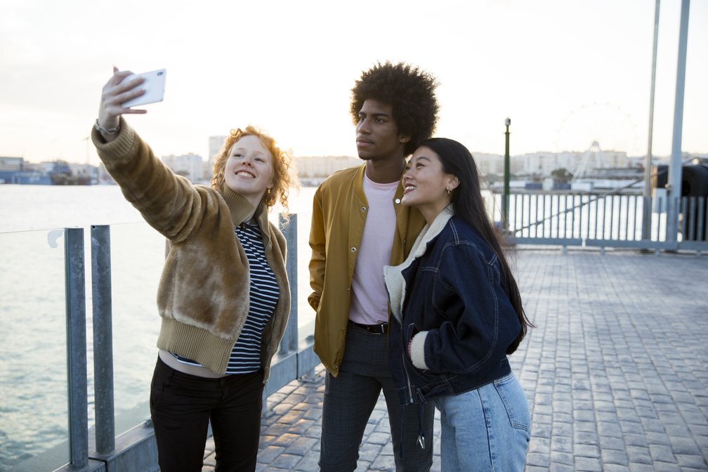 Trio de jovens viajantes tirando selfie à beira-mar em cidade internacional, representando a busca pelo melhor seguro viagem internacional para experiências seguras e inesquecíveis.